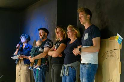 5 Personen beobachten das Schauspiel von einer Seitenwand aus; ganz links im Bild ist die Moderatorin Claudia Em zu sehen, die ins Mikrofon spricht; daneben ein Zuseher mit Baby im Arm sowie drei weitere Personen.